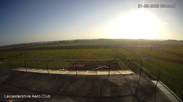 A wide view of an open grassy field under a clear sky with the sun low on the horizon, casting bright light. In the foreground, there is a concrete platform with a metal railing, and below it, an aerodrome or airfield with a runway and taxiways. The top right corner displays the date and time as "01-05-2025 06:59:50." The bottom left corner has the text "Leicestershire Aero Club."