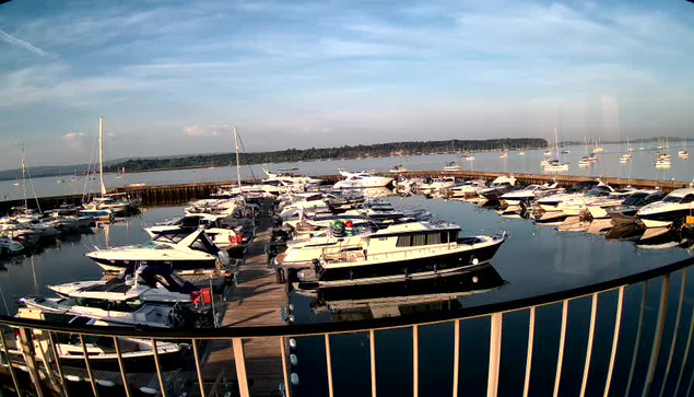 A marina filled with numerous boats and yachts docked along wooden piers. Several sailboats are anchored in the distance across the calm water, with a tree-lined shoreline under a partly cloudy sky.