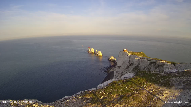 A view from a webcam showing white chalk cliffs overlooking the ocean. On top of the cliffs, there is a grassy area with a small building and some structures. In the calm sea below, there are several white rock formations and a red and white lighthouse on one of the rocks. The sky is mostly clear with a few wispy clouds. The image has a timestamp in the lower left corner and a website URL in the lower right corner.