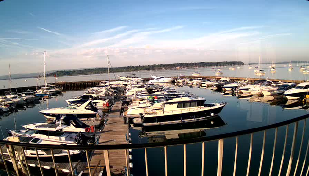 A marina filled with numerous boats docked along wooden piers extends into calm water. In the background, there are additional boats anchored farther out, with a tree-lined shoreline under a bright blue sky with wispy clouds. A metal railing is visible in the foreground.