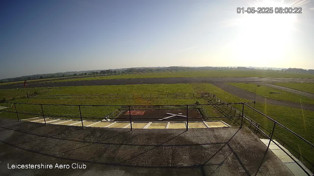 A wide view of an airfield shows a grassy landscape stretching into the distance under a clear sky, with the bright sun shining from the right side. In the foreground, there is a balcony or observation deck with a railing and some pavement. The bottom left corner has the text "Leicestershire Aero Club," and the top right corner displays the date and time as "01-05-2025 08:00:22."