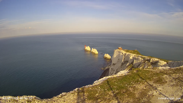 Aerial view of white chalk cliffs along a coastline, with a small building and a lighthouse on top. In the ocean below, there are several large white rock formations and a red-and-white striped lighthouse on a smaller rock island. The sky is mostly clear with a few wispy clouds. The scene is bathed in soft sunlight, creating shadows on the cliffs and rocks.