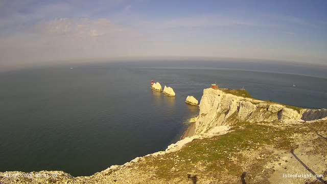 A panoramic view of white chalk cliffs overlooking the ocean under a partly cloudy sky. In the distance, there are a few boats sailing on the water, and a red and white lighthouse can be seen on one of the rocks near the coast. The foreground shows a rough, rocky path along the edge of the cliffs.