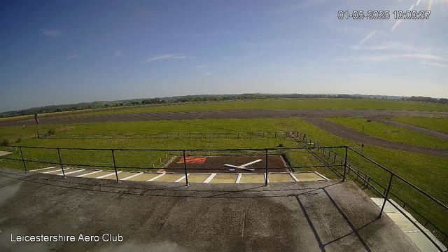 A wide view of an airfield showing a grassy runway area with taxiways and a clear blue sky overhead. There is a small parking area with white lines and a gate in the foreground, along with a safety railing. The image is timestamped "01-05-2025 10:00:37" and labeled "Leicestershire Aero Club" in the bottom left corner.