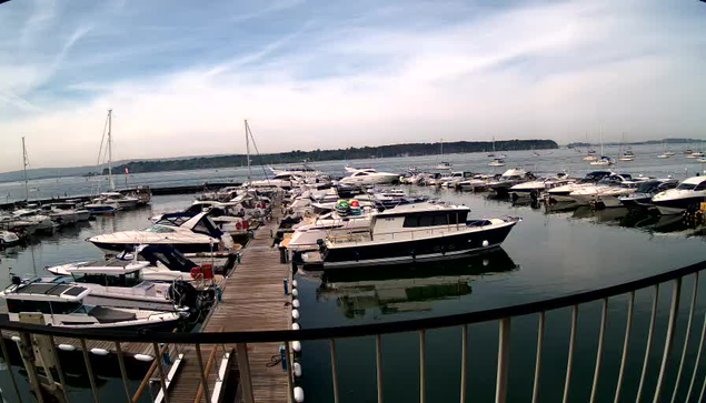 A marina with multiple boats docked along wooden piers, set against a calm body of water and a cloudy sky. The boats vary in size and style, with some featuring covered cabins and others open. The water reflects the boats and the sky, creating a peaceful scene. A metal railing is visible in the foreground.