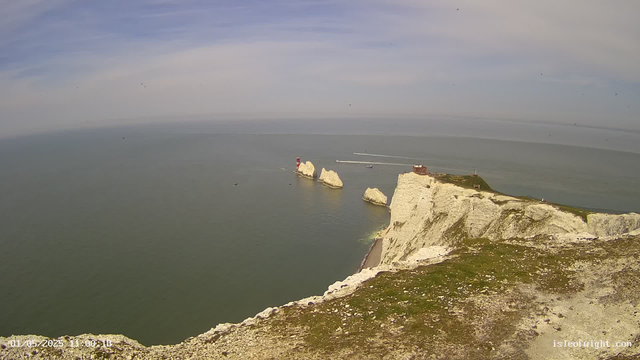 A panoramic view of white chalk cliffs along the coastline, with a lighthouse and a small building on top. The cliffs descend steeply into the calm sea, which extends to the horizon under a partly cloudy sky. Several boats are visible in the water near the cliffs. The scene is bright and appears to be taken during daylight hours.
