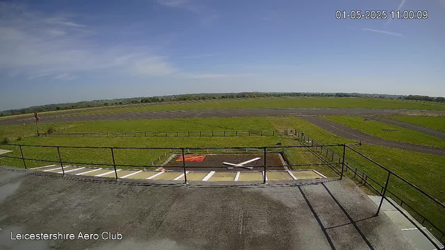 An empty parking area with a black metal railing, overlooking a grassy field and a clear blue sky. There is a red and white landing pad with a white arrow marking, and a small red sign on the left side. The scene is bright and sunny, with a few wispy clouds in the sky. The image is from a webcam at Leicestershire Aero Club, timestamped January 5, 2025, at 11:00:09.