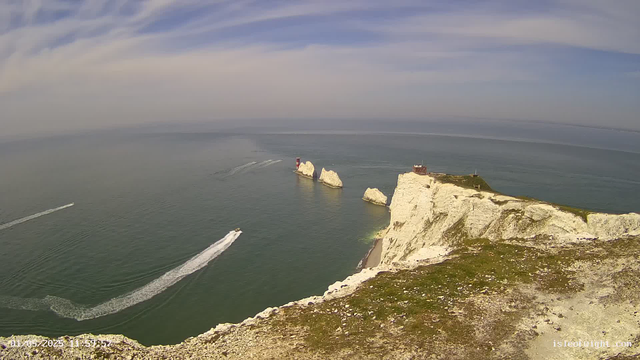 Aerial view of white chalk cliffs along the coast with three large rock stacks extending into the sea. Several boats are sailing nearby, leaving white foam trails behind. The sky is partly cloudy with patches of blue, and the ocean appears calm. There are small structures on top of the cliffs.