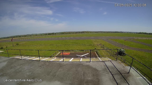 A view from a webcam at Leicestershire Aero Club showing a wide open grassy airfield with taxiways and runways in the distance. The sky is mostly clear with some scattered clouds. There are a few parked vehicles and fencing along the foreground. The timestamp in the top right corner indicates the date and time as January 5, 2025, at 12:00:08.