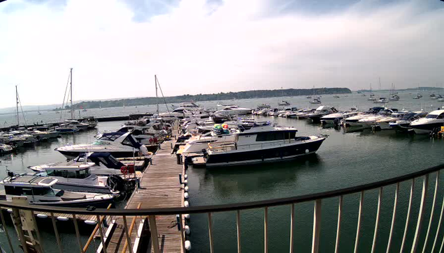 A marina with numerous boats docked along a wooden pier extending into the water. The water is calm, and the sky is partly cloudy, with a distant shoreline visible on the horizon.