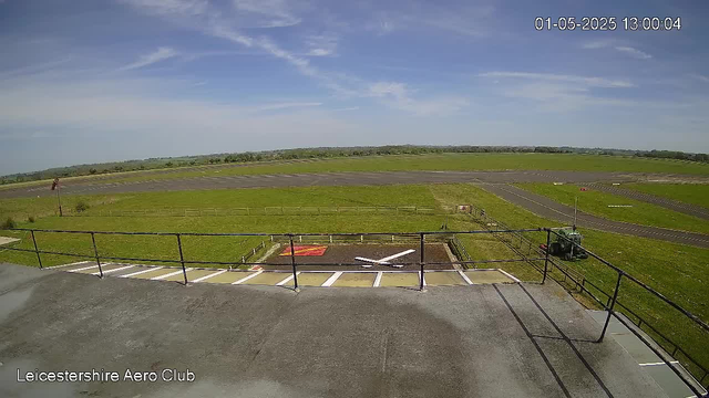 A clear view of an open grassy airfield with a runway stretching into the distance. In the foreground, there is a paved platform with a black railing, small white parking spaces, and a red marking. To the right, a small green tractor is parked near the railings. The sky is mostly blue with some scattered clouds, and the scene is bathed in bright daylight. The timestamp in the top right corner indicates the date and time as January 5, 2025, at 1:00 PM. The bottom left corner displays the text "Leicestershire Aero Club."
