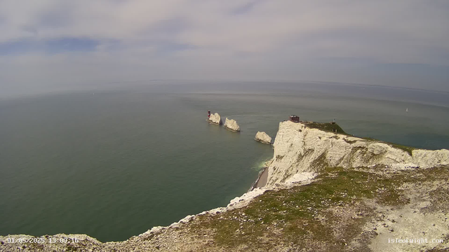 A panoramic view of white chalk cliffs extending into the sea, with a lighthouse on a small rock formation offshore. The sky is partly cloudy with patches of blue, and the ocean appears calm with a few sailboats visible in the distance. The foreground shows the rugged edge of the cliff with some grass and gravel.