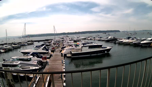 A view of a marina with multiple boats and yachts moored along a wooden dock, extending into the water. The sky is partly cloudy, and the horizon shows more boats and a distant shoreline.