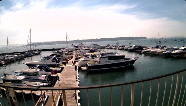 A marina with numerous boats and yachts docked along wooden piers. The water is calm, and the sky is partly cloudy with a mix of blue and white. The image is taken from a vantage point behind a curved railing. In the background, there is a landmass with some structures and trees visible in the distance.