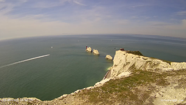 A panoramic aerial view of white chalk cliffs rising sharply from the sea, with a grassy top and a small building at the edge. Several large rock formations and a lighthouse are visible along the base of the cliffs. The ocean below is calm and dotted with sailboats and a speedboat creating a wake. The sky is partly cloudy with some wispy clouds.