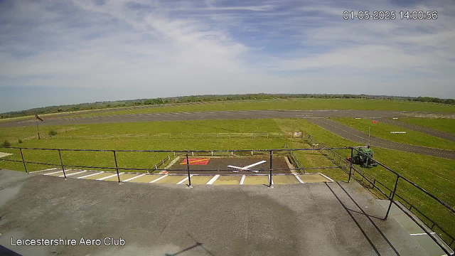 An empty aircraft runway with a small helicopter on the tarmac in the foreground. There are grassy fields surrounding the runway, and a blue sky with some clouds overhead. The image is timestamped "01-05-2025 14:00:56" and labeled "Leicestershire Aero Club" in the bottom left corner.