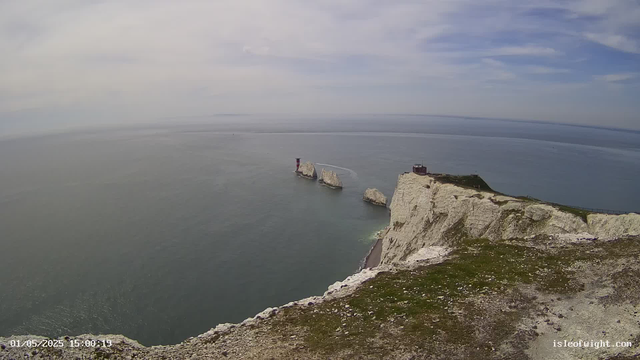 A view from a webcam shows white chalk cliffs overlooking the ocean. In the distance, there are three rock formations with a small lighthouse or tower on the middle one. The sky is partly cloudy with patches of blue, and the water appears calm. The foreground includes the edge of the cliff with some patches of grass and rocks. The timestamp indicates the date and time as May 1, 2025, at 3:00 PM.