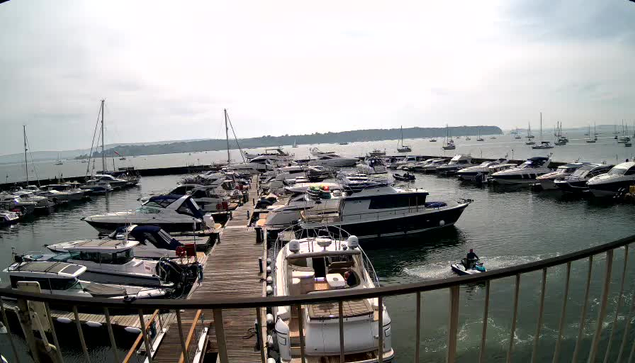 A marina filled with various boats and yachts docked along a wooden pier. Several boats are moored on either side of the pier, with some in the water nearby. In the background, there is a calm body of water extending to the horizon with more boats anchored in the distance. The sky is partly cloudy, and a landmass with trees is visible on the horizon.