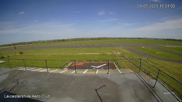 A view from a webcam at Leicestershire Aero Club showing an empty aircraft parking area with a small helipad marked by a red and yellow rectangle and a white helicopter symbol. Beyond the parking area, there is a grassy field and a runway extending into the distance under a partly cloudy blue sky. The timestamp in the top right corner indicates the date and time as January 5, 2025, at 3:01 PM.