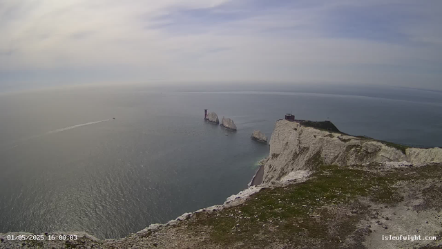 A panoramic view of white chalk cliffs extending into the sea, with large rock formations offshore and a lighthouse perched at the edge of the cliffs. The sky is partly cloudy with patches of blue.