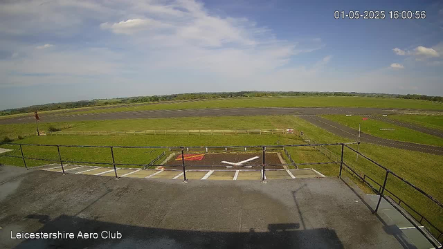 An outdoor view from a webcam at Leicestershire Aero Club showing a clear sky with a few clouds, a grassy airfield with runways in the distance, and a helicopter landing pad in the foreground. The timestamp in the top right corner reads "01-05-2025 16:00:56."