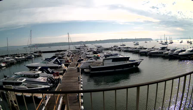 A marina with several yachts and boats docked along the pier, extending into the water. The water is calm, and there are multiple sailboats and motorboats tied to the docks. The sky is partly cloudy with patches of sunlight breaking through, creating a peaceful maritime scene. In the foreground, there's a curved railing along a balcony or walkway overlooking the marina.