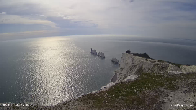 A panoramic view from a coastal cliff shows white chalk cliffs dropping sharply into the sea. Several large rock formations and sea stacks are visible in the water near the cliffs. The sky is partly cloudy with patches of sunlight reflecting off the calm ocean surface. A small building or lighthouse is situated on the edge of the cliff. The scene appears to be taken in late afternoon or early evening, as indicated by the timestamp.