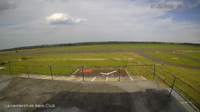 A view from a webcam showing an empty landing pad with a large white arrow painted on it, surrounded by a safety railing. Beyond the pad, there are open grassy fields and runways extending into the distance under a partly cloudy sky. The timestamp in the upper right corner reads "01-05-2025 17:00:55," and the text in the lower left corner says "Leicestershire Aero Club."
