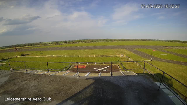 A view from a webcam showing the outside of Leicesterhire Aero Club. In the foreground, there is a rooftop with a railing and a marked helicopter landing pad with a white 'H'. Beyond the rooftop, there is a grassy area enclosed by a low fence, with a small windsock on the left. Further out, a small runway extends into the distance under a partly cloudy sky. The timestamp in the top right corner reads "01-05-2025 18:00:27."