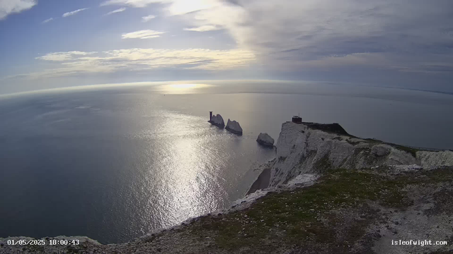 A view of white chalk cliffs along the coastline with a small building or lighthouse on top. Off the coast, there are several large sea stacks rising from the water, and the sky is partly cloudy with the sun reflecting on the calm sea below.