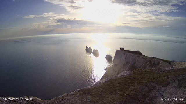 A scenic view of white chalk cliffs overlooking the ocean during sunset. The sky is partly cloudy with the sun low on the horizon, reflecting brightly on the calm sea. There are three sea stacks emerging from the water near the cliffs, and a small building is visible atop the cliffs on the right side of the image. The timestamp at the bottom left reads "01/05/2025 19:00:12," and the website "isleofwight.com" is displayed at the bottom right.