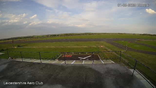 An outdoor scene showing a parking area with a small aircraft on the tarmac, surrounded by a black safety railing. Beyond the parking area, there is a vast open field with green grass and a runway curving into the distance under a partly cloudy sky. The timestamp in the top right corner reads "01-05-2025 19:00:47," and the bottom left corner displays the text "Leicestershire Aero Club."
