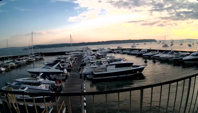 A marina at sunset with numerous boats docked along wooden piers, and a calm water surface reflecting the sky's soft colors. In the background, a distant shoreline with trees and a few more boats floating on the water. The sky is partly cloudy with a warm glow from the setting sun.