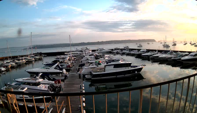 A peaceful marina filled with numerous boats and yachts docked along a wooden pier. The water reflects the boats and the sky, which is partly cloudy with soft pastel colors from the setting or rising sun. In the background, there is a distant shoreline with low hills or land extending across the horizon. A black railing curves in the foreground, framing the scene.