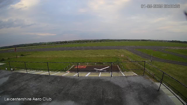 A wide view of an airfield taken from a webcam at Leicesterhire Aero Club. The foreground shows a paved surface with black metal railings and a few marked parking spaces. Beyond, there is a grassy area with a small helipad and an aircraft-shaped marker. The background features a flat landscape with a curving runway, open fields, and a partly cloudy sky. The timestamp in the top right corner reads "01-05-2025 20:00:36".