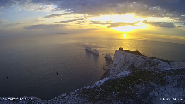A scenic view of white chalk cliffs at sunset, overlooking the ocean with a few large rock formations and a small structure on the edge of the cliff. The sky is partly cloudy, illuminated by the setting sun, casting a golden glow over the water and landscape. The image has a timestamp in the bottom left corner and the website url "isleofwight.com" in the bottom right corner.