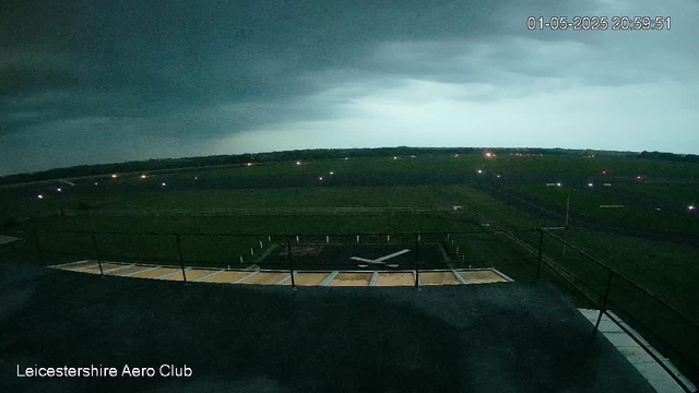A nighttime view from a webcam at Leicestershire Aero Club shows a cloudy sky with dark storm clouds and patches of rain in the distance. The horizon is faintly illuminated by city lights or lightning, creating a moody atmosphere. The scene includes a runway with a helicopter landing pad below the camera, and the surrounding open fields extend into the distance. The timestamp in the top right corner indicates the date and time as January 5th, 2025 at 8:59 PM.