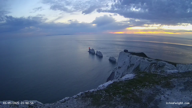 An aerial view of a coastal landscape at sunset, featuring tall white chalk cliffs with patches of greenery, a lighthouse on one of the cliffs, and several large rock formations in the calm sea. The sky is partly cloudy with the sun setting on the horizon, casting a warm glow. The timestamp in the bottom left corner reads "01/05/2025 21:00:56," and the bottom right corner displays the website "isleofwight.com."