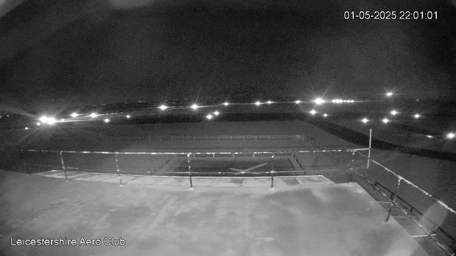 A black-and-white nighttime image of a sports field viewed from a webcam at Leicesterhire Aero Club. The field is enclosed by a low fence, with a small goalpost visible in the center. In the distance, there are several bright lights scattered across the horizon, illuminating the dark sky. The top right corner shows the date and time as "01-05-2025 22:01:01."