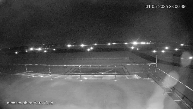 A black-and-white image from a webcam showing an empty aircraft parking area at night. There are a few lights in the distance illuminating the horizon, and faint reflections on the tarmac. The timestamp indicates it was taken on January 5th, 2025, at 11:00 PM. The bottom left corner has text that reads "Leicestershire Aero Club."