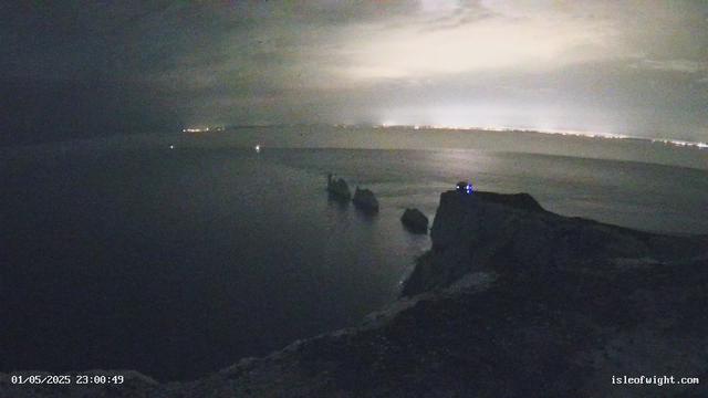 A dark nighttime scene of a beach with large rocks in the foreground and calm ocean waters stretching into the distance. The horizon is faintly illuminated by city lights, creating a soft glow against the cloudy sky. There is a tiny, illuminated structure or light on one of the rocks near the right side of the image. The timestamp in the lower left corner reads "01/05/2025 23:00:49," and the bottom right corner has the website "isleofwight.com."