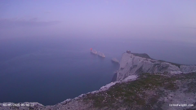 A view from a cliff overlooking the ocean at dawn or dusk. The sky is a soft gradient of purple and pink, with faint clouds. In the distance, there are two small boats or ships on the water, one with a red light. The foreground shows the edge of the white chalk cliff with some grassy patches and rocky terrain. The timestamp in the lower left corner indicates "02/05/2025 05:00:22," and the website "isleofwight.com" is visible in the lower right corner.