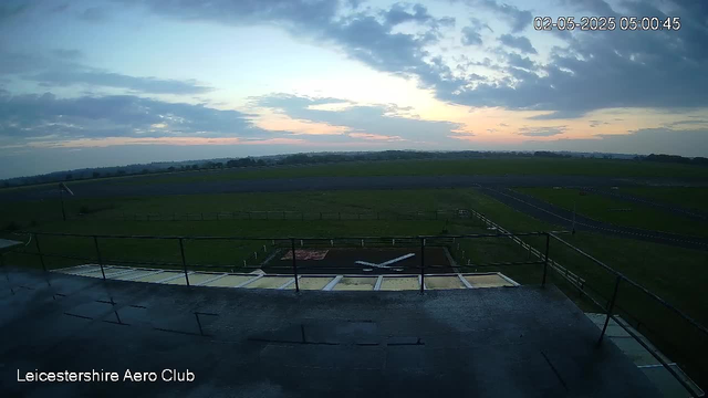 A view from a webcam showing an airfield during dawn, with a small airplane parked on a platform in the foreground. The sky is partly cloudy with the sun beginning to rise, casting a soft light over the green grass and runway in the distance. The image is timestamped "02-05-2025 05:00:45" and It is labeled "Leicestershire Aero Club" in the bottom left corner.