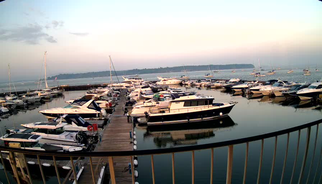 A calm marina filled with numerous boats and yachts docked along wooden piers, with some boats reflected on the still water. The sky is lightly cloudy with a soft gradient of colors as the sun sets or rises, creating a serene and peaceful atmosphere. A railing runs along the foreground, curving with the edge of the marina.
