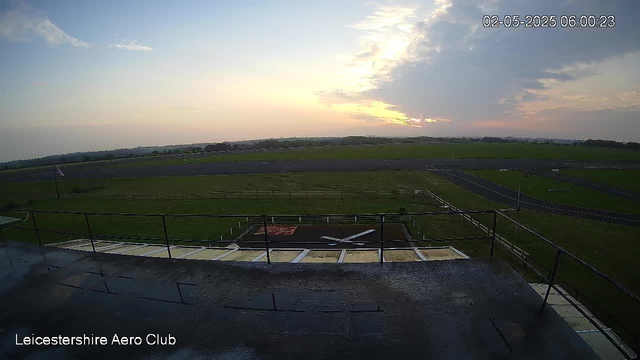 A wide view of an open grassy field and a runway beneath a partly cloudy sky during sunset. The image is from a webcam at Leicestershire Aero Club, dated February 5, 2025, at 6:00 AM. In the foreground, there is a flat rooftop with a railing, and on the ground below, there is a small model airplane and some painted markings.
