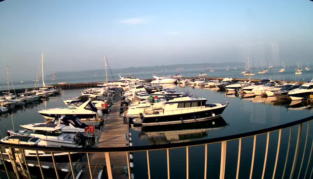 A marina filled with numerous white and dark-colored boats docked along wooden piers. The water is calm, reflecting the boats and the light blue sky above. In the background, there are more boats floating further out on the water, with a distant shoreline visible on the horizon. A curved railing is in the foreground.