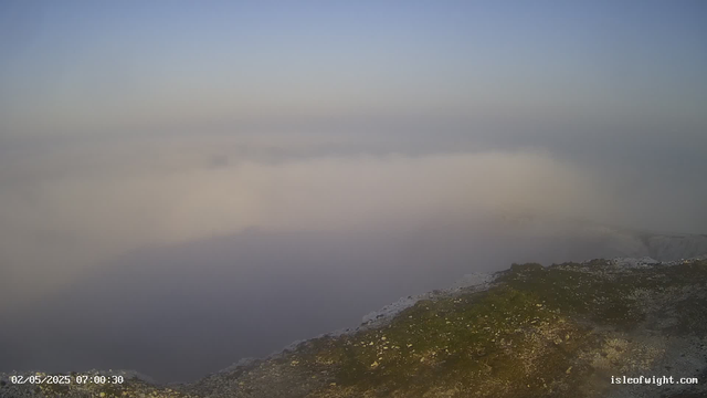 A view from a webcam showing a foggy or cloudy landscape with a rocky, moss-covered foreground and a thick layer of fog or low clouds stretching across the scene. The sky above is clear with a faint gradient from blue at the top to lighter near the horizon. There is a timestamp in the lower left corner showing "02/05/2025 07:00:30" and a website URL "isleofwight.com" in the lower right corner.