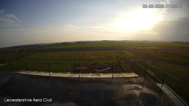 An outdoor scene at the Leicesterhire Aero Club showing a grassy airfield under a bright, partly cloudy sky with the sun low on the horizon. There are some walkways and railings in the foreground, and distant rolling hills stretch across the background. The image is captured during early morning hours.