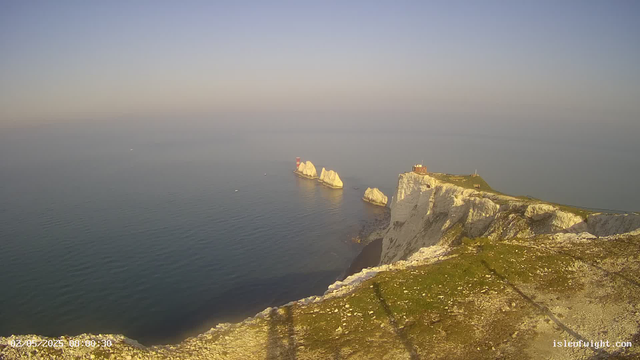 A panoramic view of white chalk cliffs overlooking the calm ocean during sunrise or sunset. The cliffs extend from the right side into the distance, with a small building or structure at the edge, and large rock formations emerging from the water near the cliffs. The sky is clear with a soft gradient from light to darker blue. The water below is gently rippled, reflecting the sky's colors.