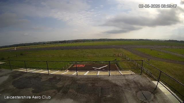 A wide view of an open grassy area with a small helipad marked with a white 'H' in the foreground. There are parking spaces with white lines, some of which are empty, and a black metal railing surrounding the area. The landscape extends into rolling fields and a distant tree line under a partly cloudy sky. The timestamp in the top right corner reads "02-05-2025 08:59:47," and there is text in the bottom left corner that says "Leicestershire Aero Club."